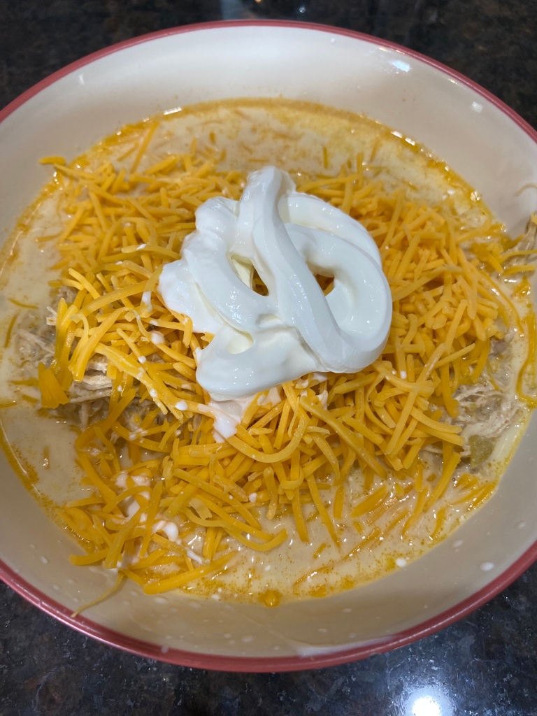 White Chicken Chili topped with shredded American cheese and sour cream in a bowl with white inside and maroon colored outside. 